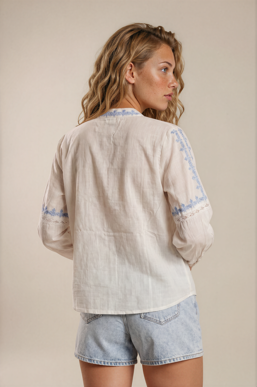 Close up image of model wearing white boho top with 3/4th sleeves and blue embroidery. This white button-down top is perfect for the summer. The sleeves have a beautiful lace inserts and the front has pleats. 